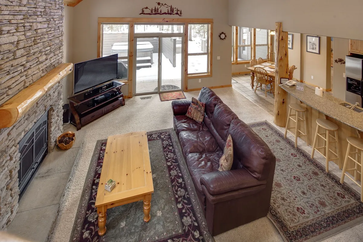 A spacious open-concept living area featuring a large brown leather sofa, a stone-faced gas fireplace, wooden coffee table, kitchen bar area with stools, and a dining space with wooden chairs. Large windows flood the room with natural light, and the seamless flow between living, kitchen, and dining encourages group interaction.