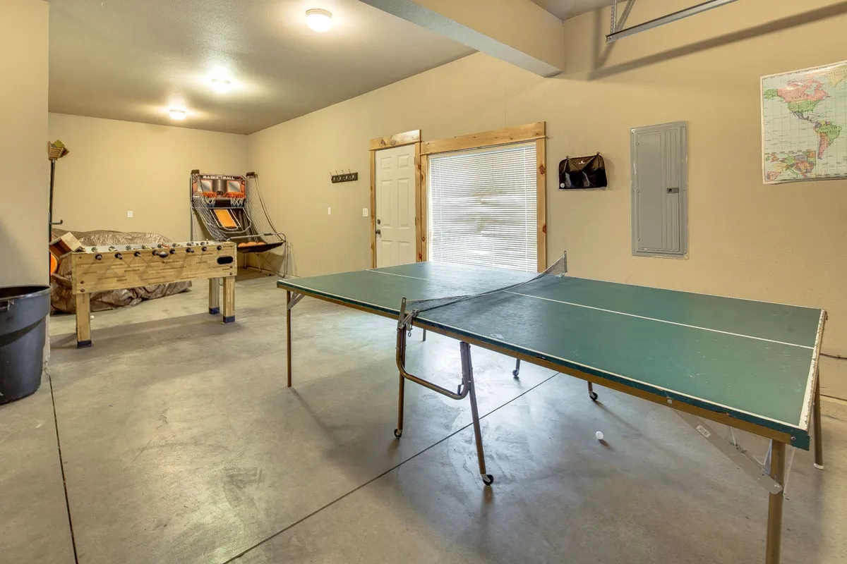 The lively garage game room is front and center, featuring both a ping pong table and a foosball table as the main attractions. The space is energetic and welcoming, with a door leading outside that hints at access to bikes and further adventure. The relaxed, playful mood is enhanced by imagined scenes of friends and family laughing and competing.