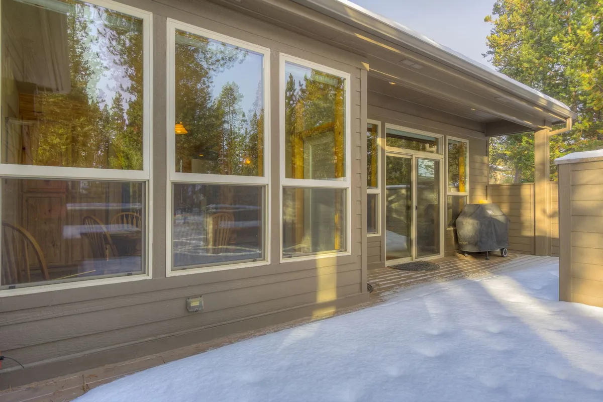 This exterior shot captures the home's inviting patio area, framed by large windows and a snow-covered landscape. The grill hints at outdoor dining possibilities, while the setting emphasizes the peaceful Sunriver surroundings and connection to nature.
