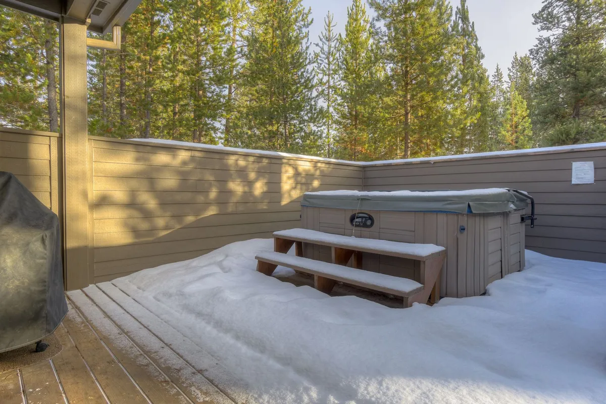 A serene outdoor hot tub set on wooden steps, surrounded by snowy ground and tall pine trees. The setting offers a glimpse of relaxation after a day of adventure, blending comfort with the natural environment.