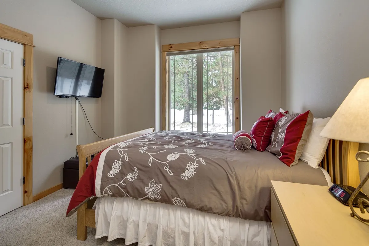 A serene queen bedroom with a bed adorned with decorative pillows, a large window overlooking the forest, and a nightstand with a lamp. The soothing color palette and natural light create a peaceful retreat for guests.