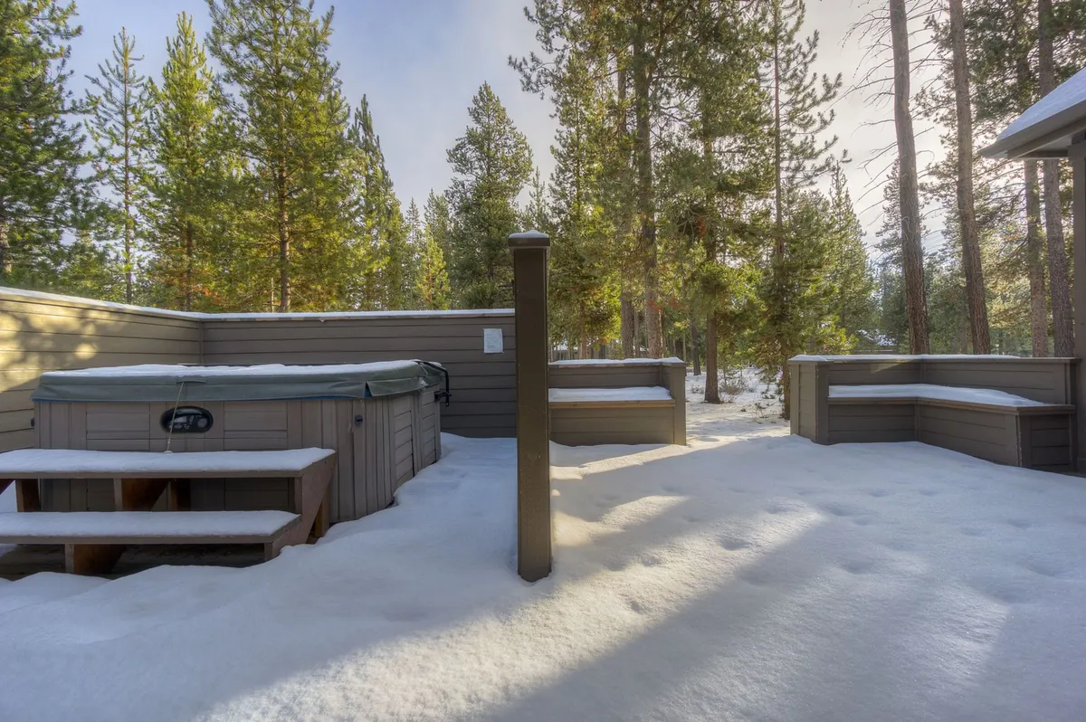 A scenic view of the hot tub nestled among tall pines, snow blanketing the deck and landscape. The setting is tranquil, perfect for winter relaxation, and highlights the allure of enjoying a soak while surrounded by nature and seasonal beauty.