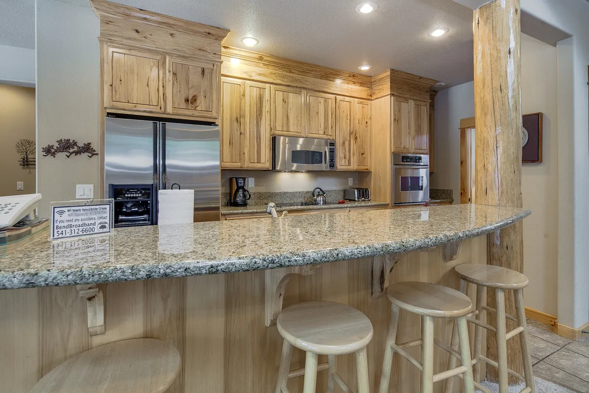 A warm, inviting kitchen with granite countertops, handsome wood cabinetry, and bar stools lined up at the counter. Modern appliances are visible, and the space is clearly designed for both cooking and socializing.