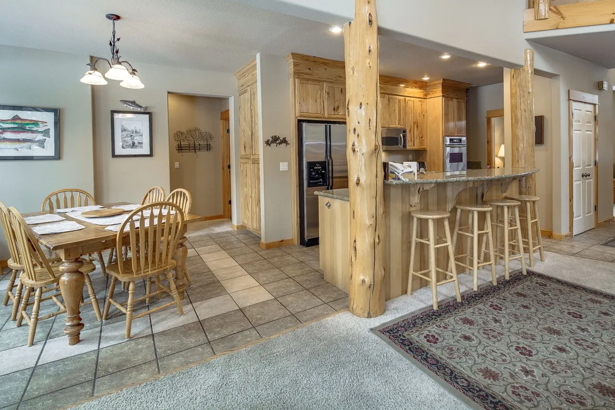 A cozy dining area with a wooden table and chairs, visible kitchen counter with stools, and rustic wooden beams overhead. The space feels communal and is bathed in natural light, inviting shared meals and laughter.