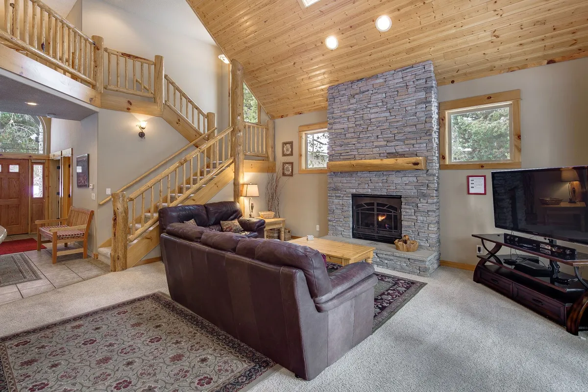 A warm, inviting interior living space featuring a striking stone fireplace, leather sofa, and large windows that let in natural light. The wooden staircase and spacious layout set a scene perfect for cozy relaxation after a day of adventure.