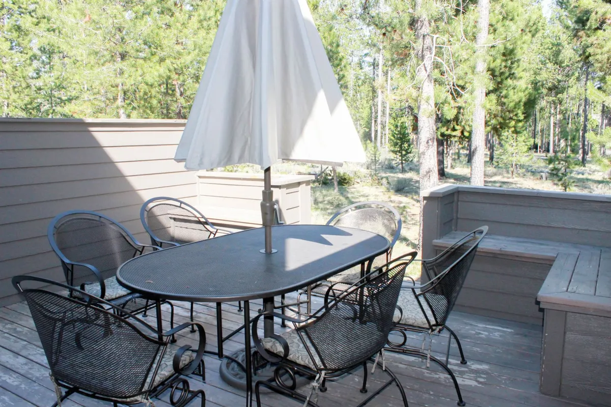 This exterior shot captures a black oval dining table with six chairs and a white umbrella on a wooden deck, surrounded by trees. The space feels relaxed and sociable, ideal for outdoor meals, reading, or celebrating with friends and family in a resort-like setting.